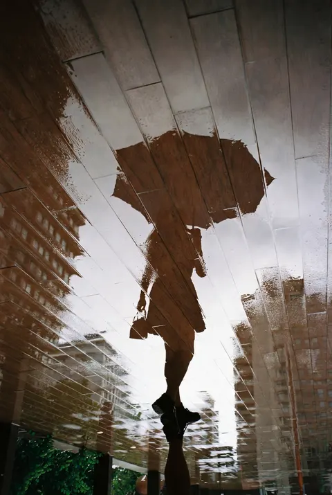 手机实拍照片，特殊倒置视角，画面主体是雨后湿漉漉的深棕色木质地板反光倒影，人物倒影和城市建筑全部倒映在有水的地板上。
倒影里：一位女生侧身背影，看不见脸部，长发，身穿深色短裙，手里举着一把大伞，另一只手拎着小皮包，穿着黑色中筒袜与亮面小皮鞋。倒影中同时倒映出阴天云层、多座城市摩天高楼。画面左下角露出真实场景的绿色灌木丛。整体是阴雨天柔和漫射自然光，画面只有倒影是视觉主体，真实人物不在画面内，只有镜面反射影像。胶片日常抓拍质感，色调清冷柔和，画面略带湿润朦胧感。竖版构图。