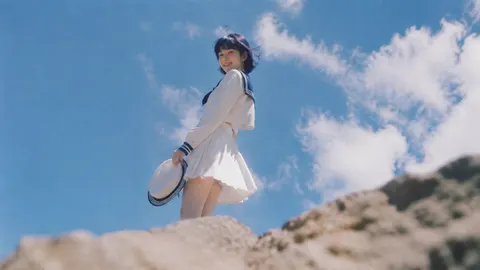 Prompt: Showa 1980s Japanese retro cinematic outdoor capture, an elegant and slender East Asian girl stands on a rough rock, with an imperceptible edge contour light on her body, ultra-low wide-angle telephoto lens, dramatic back shot from bottom to top, and the character is set off by the vast blue sky and soft white clouds. The girl has a cold fox-like facial features, a small V-long face, cold white and transparent skin, jewel blue oil bright Lobo retro short hair, and the short hair is blown by the summer breeze. Wearing a white sailor collar long-sleeved uniform, navy stripe decoration, a white pleated skirt, holding a round sailor hat in one hand in one hand, gently triggering the silk in the other hand, bowing his head to show a bright and natural smile to the camera, the corners of his mouth slightly open, and smiling with crescent eyes.    Real outdoor photography, FUJIFILM X100VI, 35mm lens, f/2.8, classic chrome simulation, Kodak Portra 160 color palette, medium-frame glue Aesthetics, low saturation cold shade, strong midday sunlight from above the side, white clothing and skin slightly overexposed, soft highlight overflow, hard geometric shadow.    The foreground rock is strongly blurred, ultra-long-angle lens capture, the background blue sky fades, shallow depth of field, slight hand-held shaking, dynamic shadow dragging, old-fashioned CCD low-resolution texture, fine film particles, real optical defects, misty dream core atmosphere, poetic realism, instant capture feeling, slightly unbalanced composition, movie theater snapshot , 9:16 vertical width.    Negative words:    Close up, portrait crop, studio lighting, artificial skin, plastic face, anime style, CGI, 3D render , extra fingers, distorted hands, exaggerated smile, oversaturated colors, modern fashion, messy bac Kground, text, logo